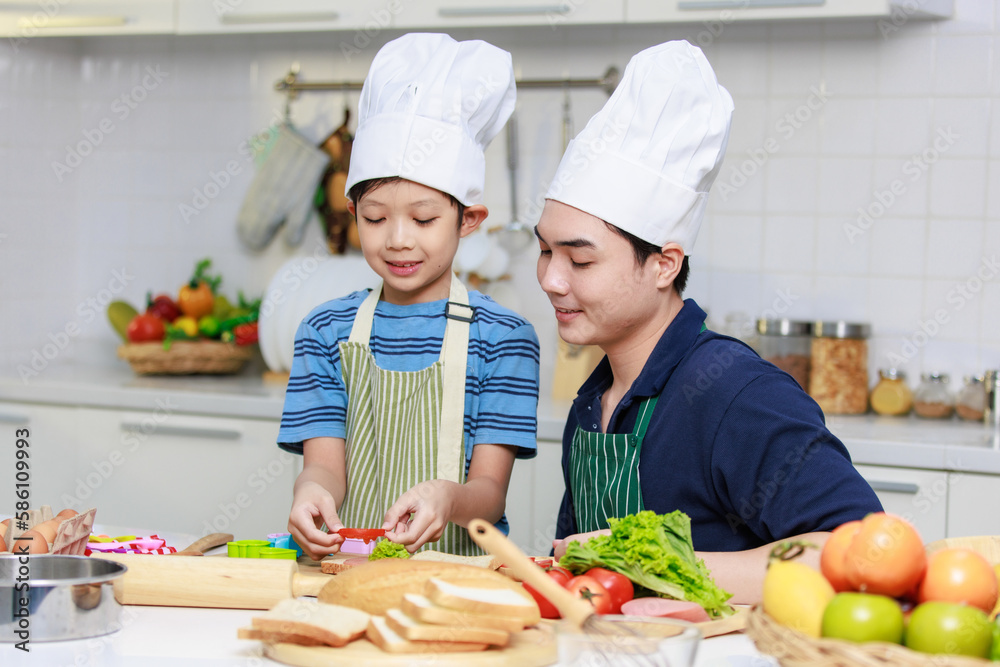 Asian young little boy chef wearing white tall cook hat and apron standing smiling holding putting sliced tomato on bread making preparing sandwiches while father helping teaching in home kitchen Asian young little boy chef wearing white tall cook hat and apron standing smiling holding putting sliced tomato on bread making preparing sandwiches while father helping teaching in home kitchen