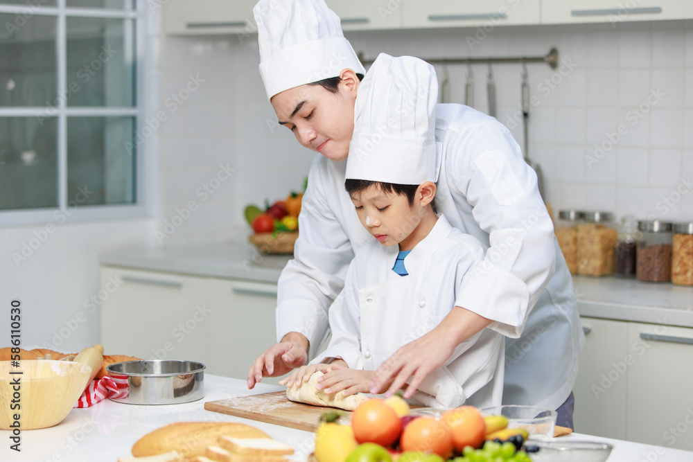 Asian young little boy chef in uniform with white tall cook hat standing learning preparing massaging flour dough on wooden cutting board while male cooking teacher helping teaching in home kitchen Asian young little boy chef in uniform with white tall cook hat standing learning preparing massaging flour dough on wooden cutting board while male cooking teacher helping teaching in home kitchen
