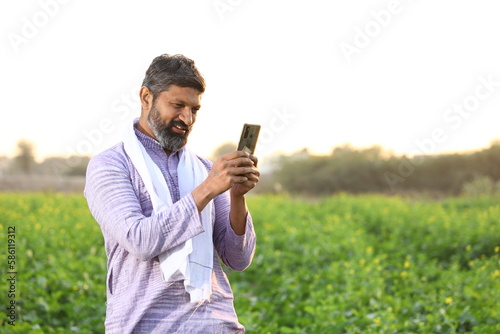 Happy rural Indian farmer chatting on his smart phone while standing in a mustard field. 