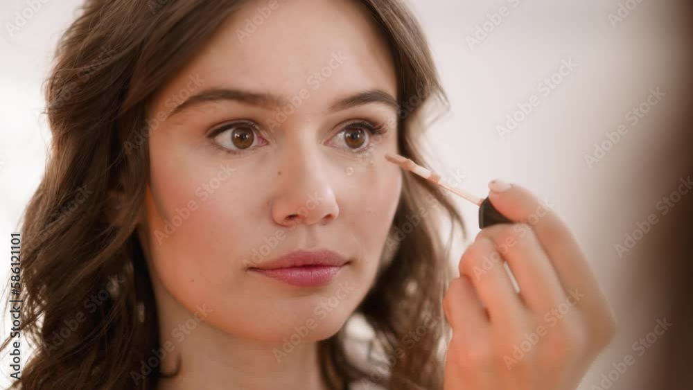 Lady applies beige concealer on skin under eyes with applicator looking in mirror. Young woman uses cosmetic product on blurred background closeup Lady applies beige concealer on skin under eyes with applicator looking in mirror. Young woman uses cosmetic product on blurred background closeup