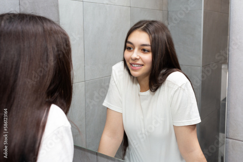 Smiling young girl looks in the mirror. Girl with long dark hair in the bathroom looks at herself in the mirror.