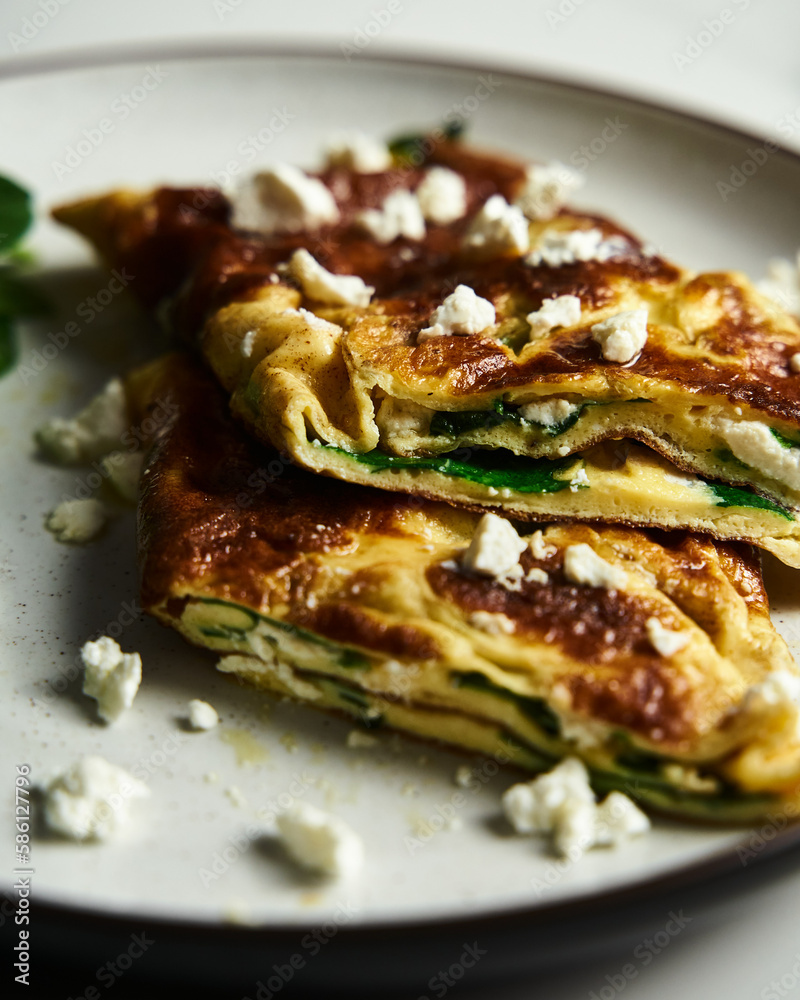 Close up of sliced folded omelette stuffed with spinach and feta on the white plate with spinach leaves on back. Healthy and dietary breakfast dish. Minimalistic food. White marble background. Close up of sliced folded omelette stuffed with spinach and feta on the white plate with spinach leaves on back. Healthy and dietary breakfast dish. Minimalistic food. White marble background.