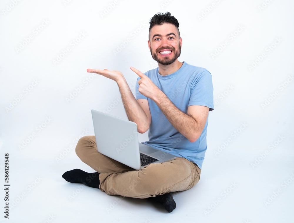 Young caucasian man with laptop sitting over white studio pointing and holding hand showing adverts Young caucasian man with laptop sitting over white studio pointing and holding hand showing adverts