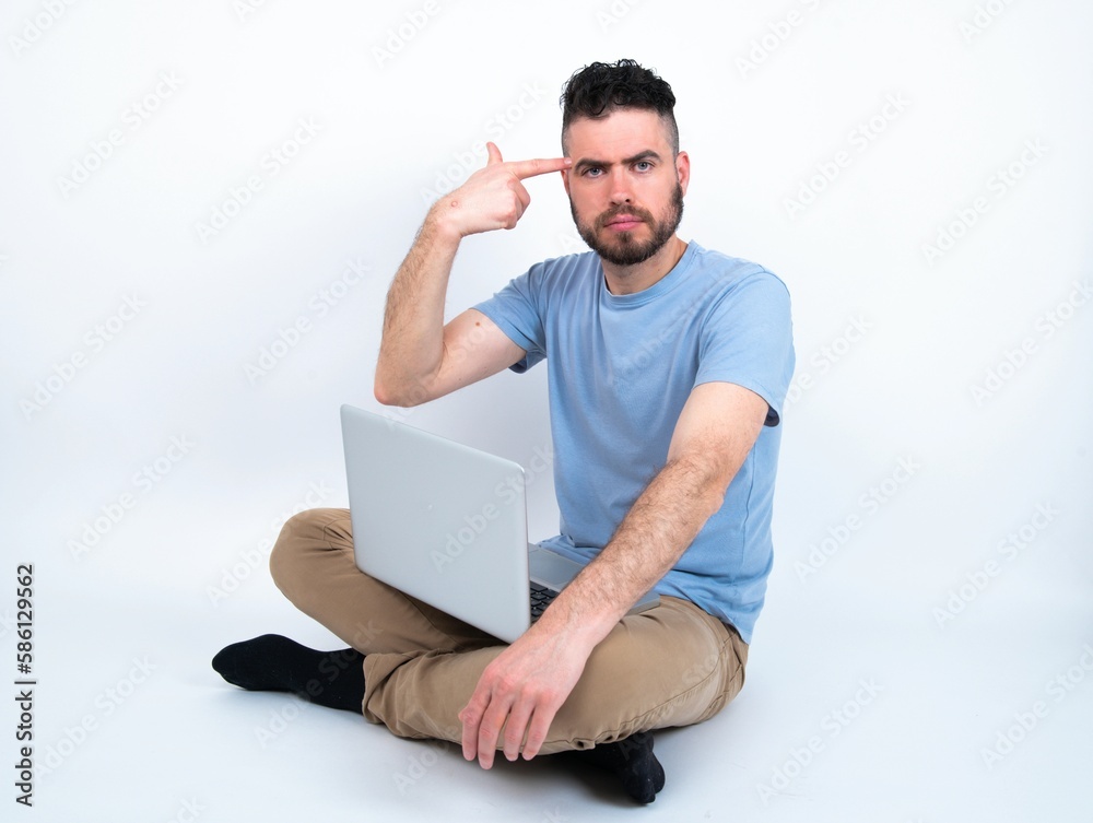 Unhappy Young caucasian man with laptop sitting over white studio makes suicide gesture and imitates gun with hand, curves lips keeps two fingers on temple, shoots, being tired of everything, Unhappy Young caucasian man with laptop sitting over white studio makes suicide gesture and imitates gun with hand, curves lips keeps two fingers on temple, shoots, being tired of everything,