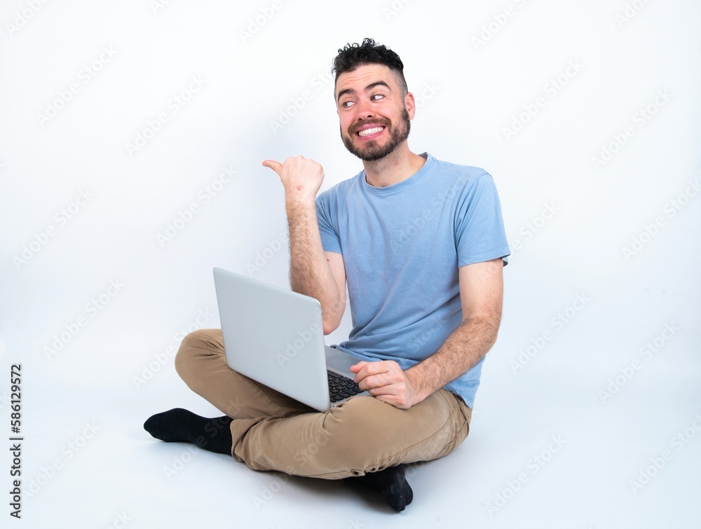 Charming Young caucasian man with laptop sitting over white studio looking at copy space having advertisements Charming Young caucasian man with laptop sitting over white studio looking at copy space having advertisements