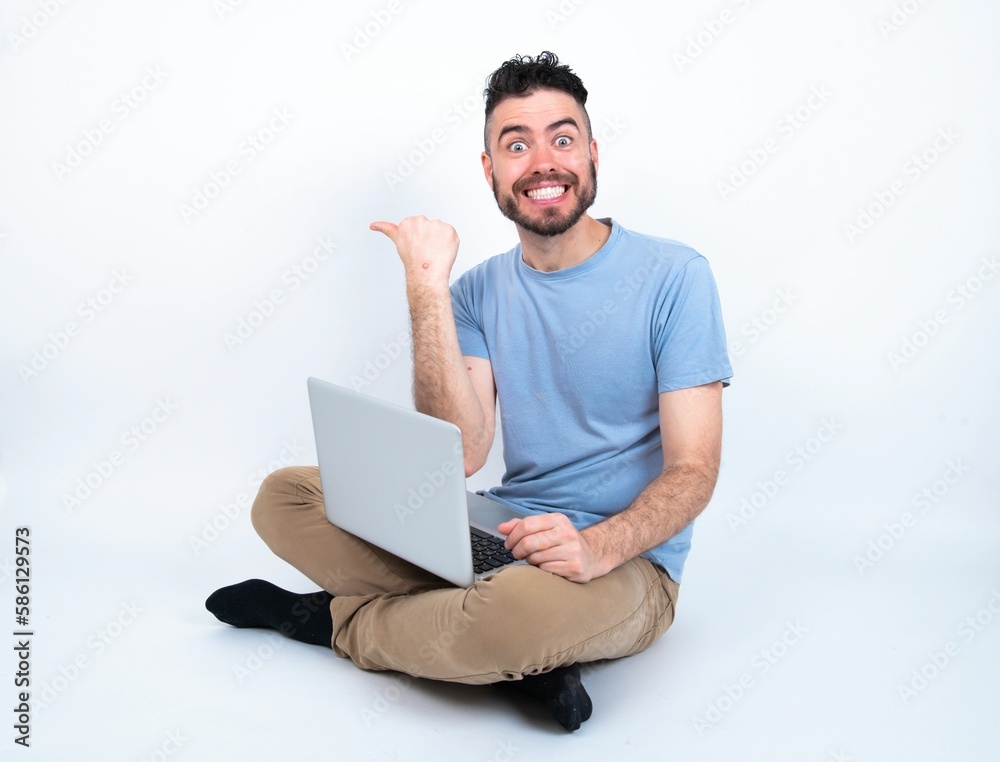 Lovely Young caucasian man with laptop sitting over white studio pointing aside with forefinger, showing at copy space having news about bargains