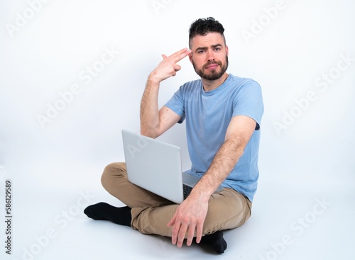 Unhappy Young caucasian man with laptop sitting over white studio makes suicide gesture and imitates gun with hand, curves lips, keeps two fingers on temple