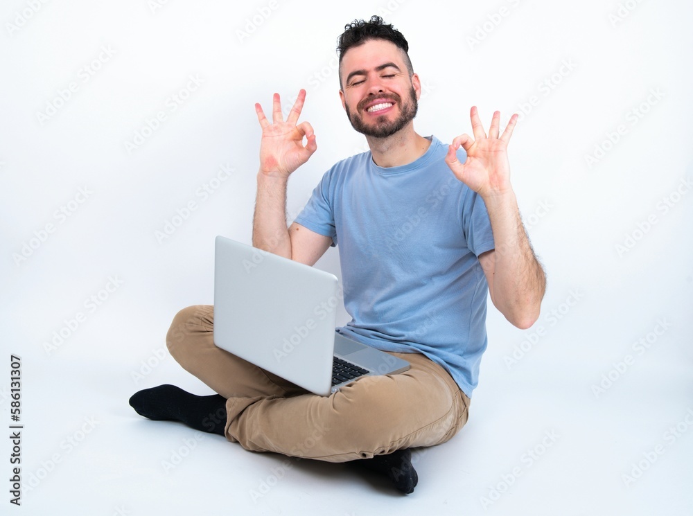 Young caucasian man with laptop sitting over white studio relax and smiling with eyes closed doing meditation gesture with fingers. Yoga concept.