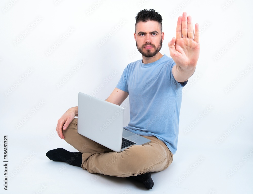 Young caucasian man with laptop sitting over white studio doing stop sing with palm of the hand. Warning expression with negative and serious gesture on the face. Young caucasian man with laptop sitting over white studio doing stop sing with palm of the hand. Warning expression with negative and serious gesture on the face.