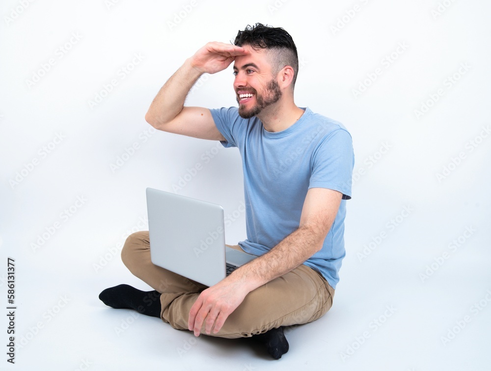 Young caucasian man with laptop sitting over white studio very happy and smiling looking far away with hand over head. Searching concept. Young caucasian man with laptop sitting over white studio very happy and smiling looking far away with hand over head. Searching concept.