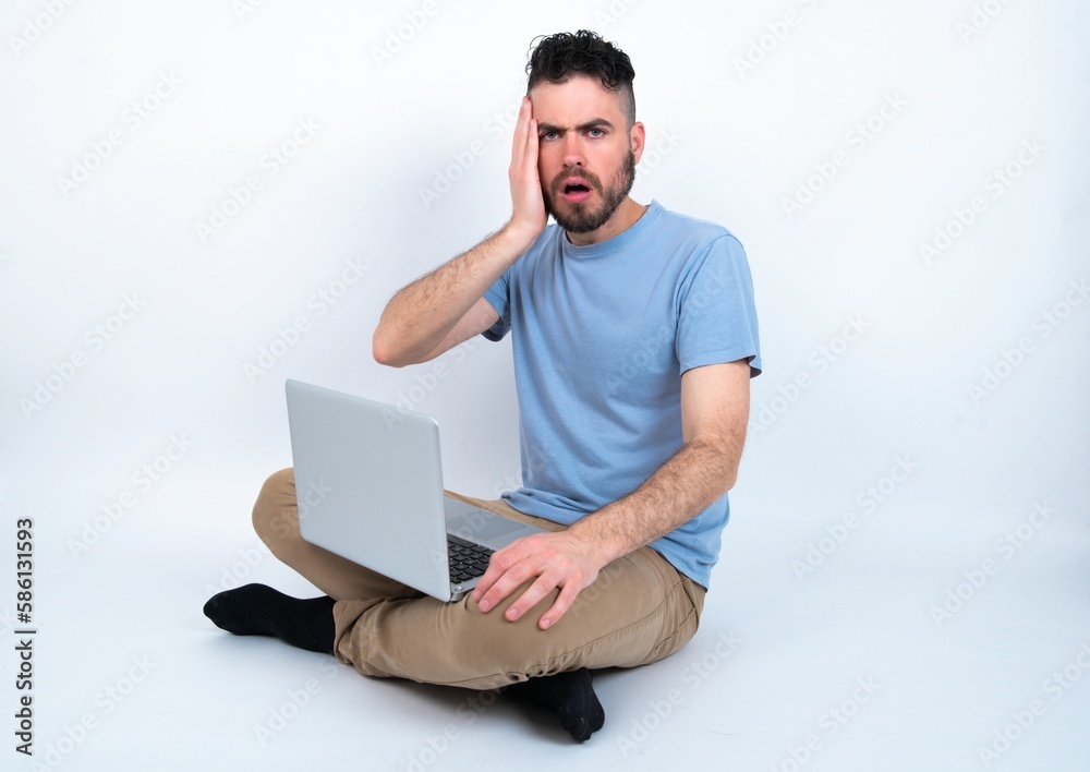 Embarrassed Young caucasian man with laptop sitting over white studio with shocked expression, expresses great amazement, Puzzled model poses indoor