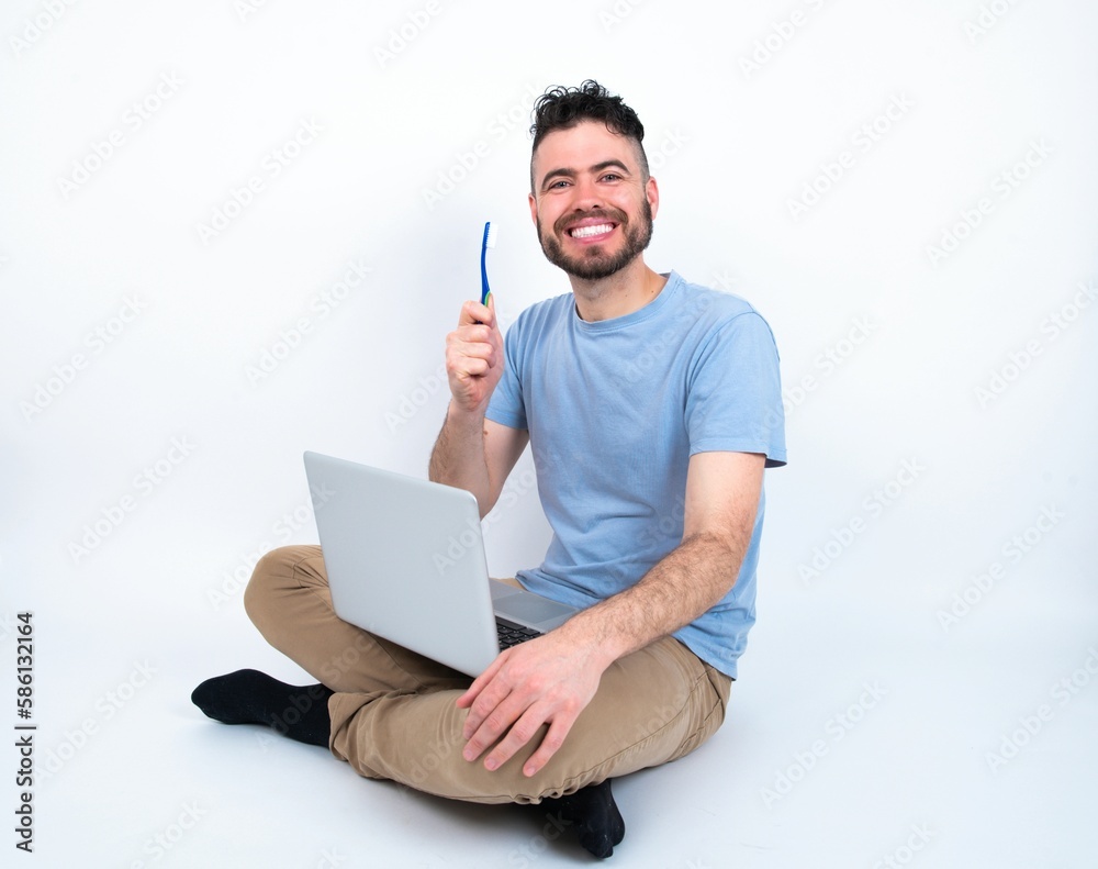 Young caucasian man with laptop sitting over white studio holding a toothbrush and smiling. Dental healthcare concept. Young caucasian man with laptop sitting over white studio holding a toothbrush and smiling. Dental healthcare concept.