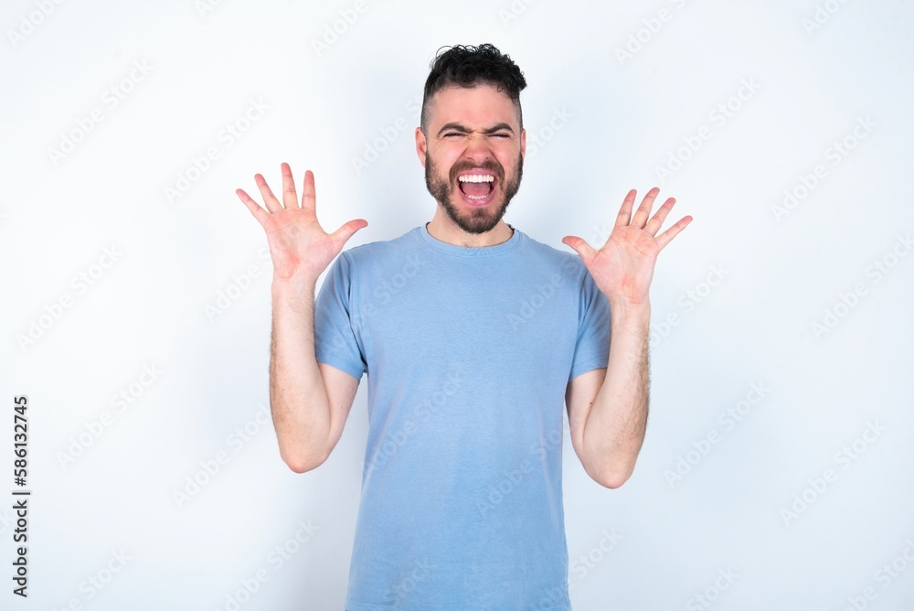 Crazy outraged Young caucasian man wearing blue T-shirt over white background screams loudly and gestures angrily yells furiously. Negative human emotions feelings concept Crazy outraged Young caucasian man wearing blue T-shirt over white background screams loudly and gestures angrily yells furiously. Negative human emotions feelings concept