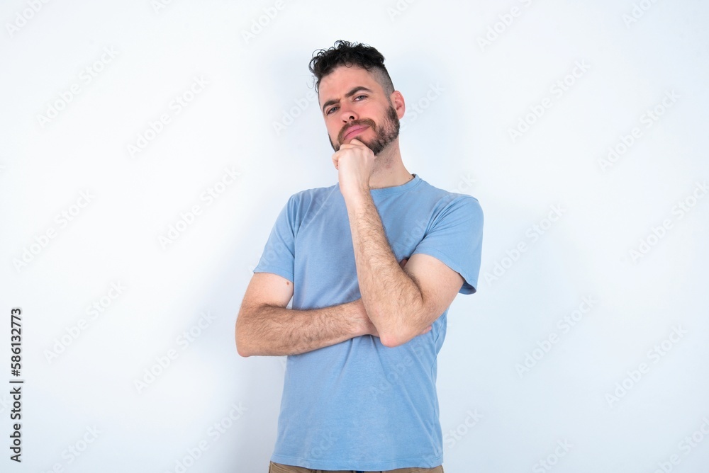Young caucasian man wearing blue T-shirt over white background looking confident at the camera smiling with crossed arms and hand raised on chin. Thinking positive.