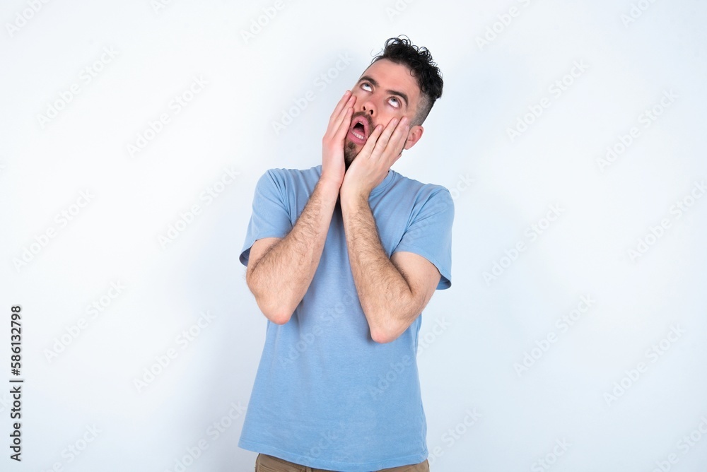 Young caucasian man wearing blue T-shirt over white background keeps hands on cheeks has bored displeased expression. Stressed hopeless model Young caucasian man wearing blue T-shirt over white background keeps hands on cheeks has bored displeased expression. Stressed hopeless model