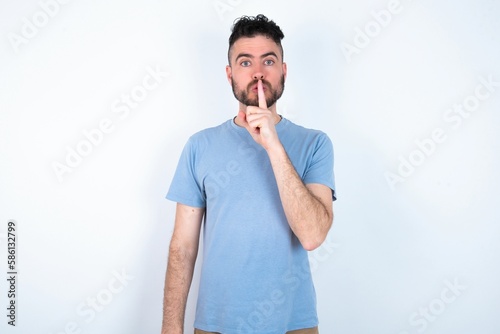Surprised Young caucasian man wearing blue T-shirt over white background makes silence gesture, keeps finger over lips and looks mysterious at camera