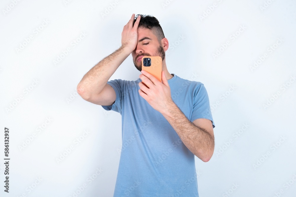 Upset depressed Young caucasian man wearing blue T-shirt over white background makes face palm as forgot about something important holds mobile phone expresses sorrow and regret blames Upset depressed Young caucasian man wearing blue T-shirt over white background makes face palm as forgot about something important holds mobile phone expresses sorrow and regret blames