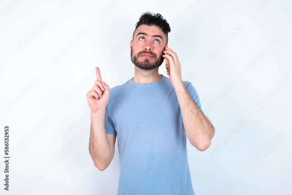 Young caucasian man wearing blue T-shirt over white background speaks on mobile phone spends free time indoors calls to friend. Young caucasian man wearing blue T-shirt over white background speaks on mobile phone spends free time indoors calls to friend.