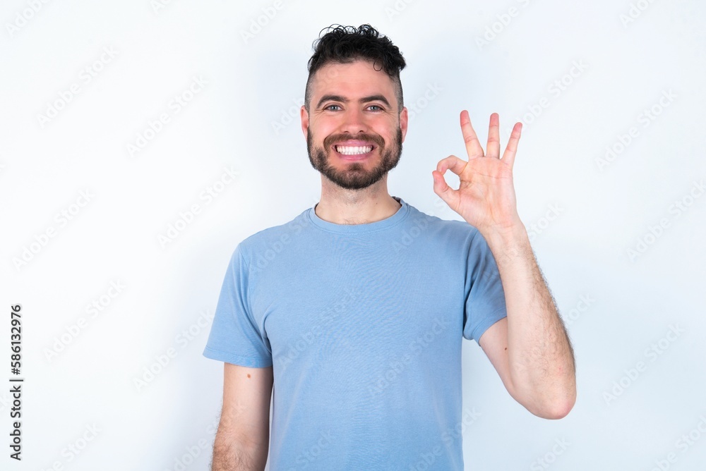 Young caucasian man wearing blue T-shirt over white background hold hand arm okey symbol toothy approve advising novelty news