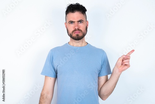 Serious Young caucasian man wearing blue T-shirt over white background smirks face points away on copy space shows something unpleasant. Look at this advertisement. Big price concept