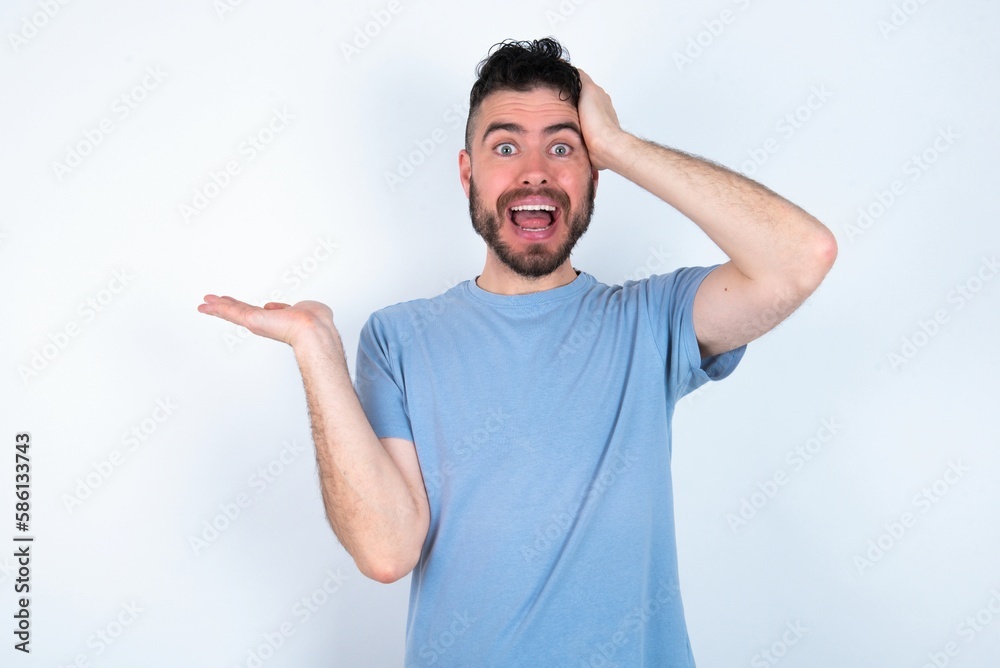 Shocked amazed surprised Young caucasian man wearing blue T-shirt over white background hold hand offering proposition Shocked amazed surprised Young caucasian man wearing blue T-shirt over white background hold hand offering proposition