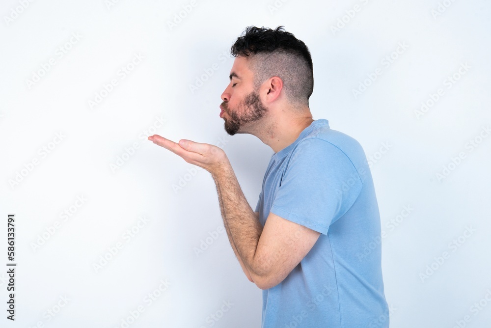 Profile side view, portrait of attractive Young caucasian man wearing blue T-shirt over white background sending air kiss