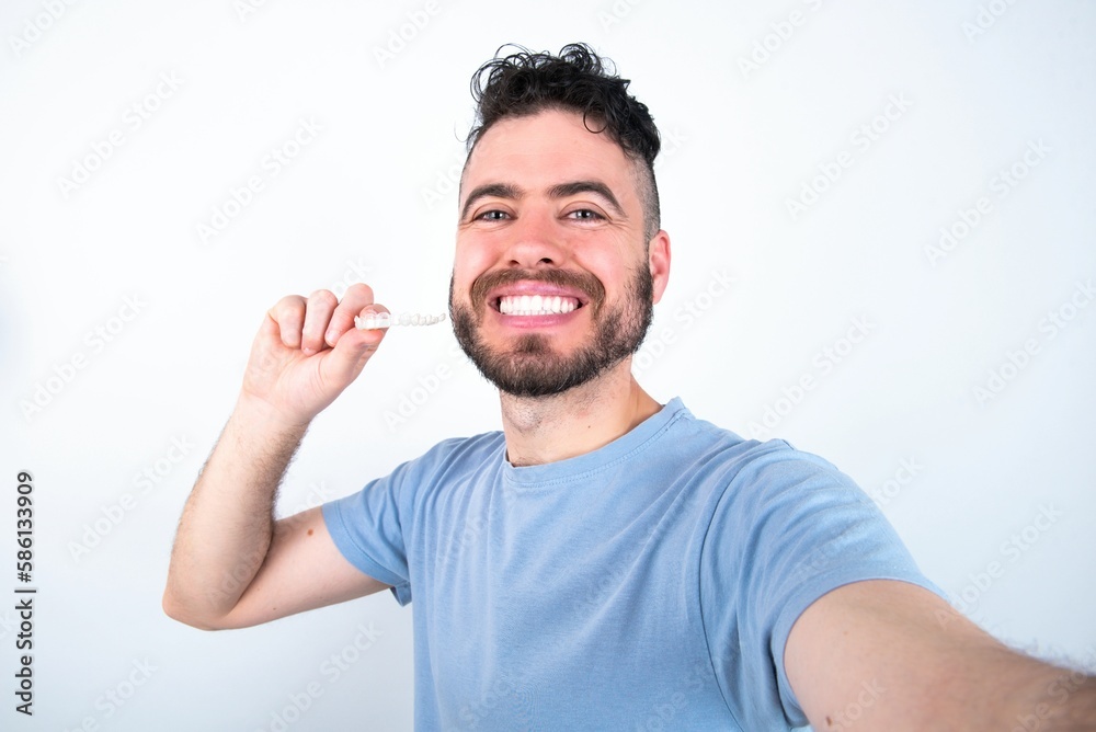 Fototapeta premium Young caucasian man wearing blue T-shirt over white background make selfie holding an invisible braces aligner, recommending this new treatment. Dental healthcare concept.