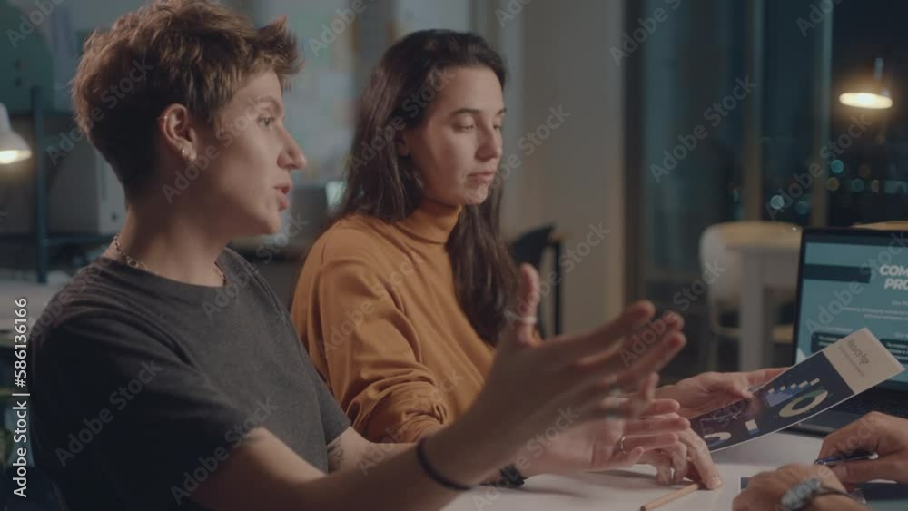 Two young businesswomen discussing papers and presentation on laptop with male colleague, having a team meeting in the office during night time Two young businesswomen discussing papers and presentation on laptop with male colleague, having a team meeting in the office during night time
