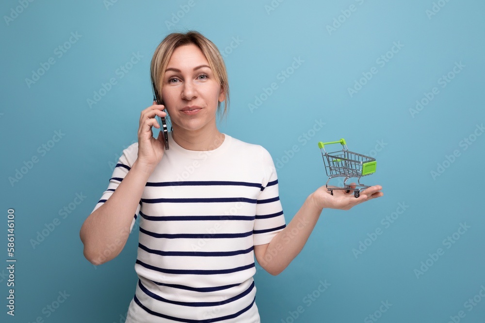 horizontal photo of attractive shopaholic girl talking on phone holding shopping cart on blue background with copy space horizontal photo of attractive shopaholic girl talking on phone holding shopping cart on blue background with copy space