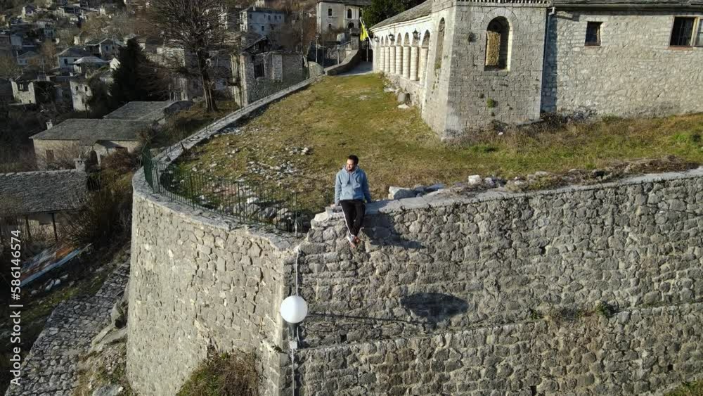 Flying backwards away from a young guy sitting in front of a Greek church bell tower in a mountain landscape village Flying backwards away from a young guy sitting in front of a Greek church bell tower in a mountain landscape village