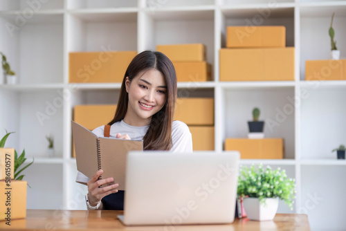 Happy young Asian woman entrepreneur, Take note after check order and Smile for sale success of online shopping store, Concept of the merchant, small business, online business, and eCommerce. © wichayada