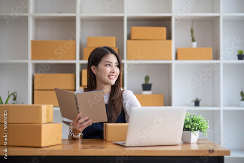 Happy young Asian woman entrepreneur, Take note after check order and Smile for sale success of online shopping store, Concept of the merchant, small business, online business, and eCommerce. © wichayada