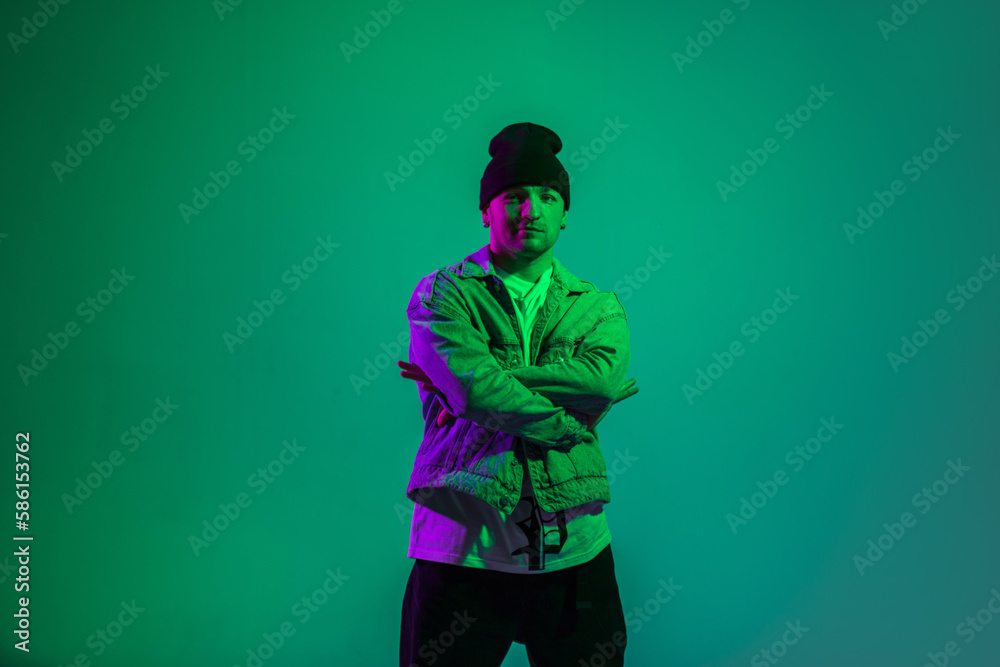 Stylish handsome trendy b-boy dancer man in trendy fashion denim clothes with a hat and jeans jacket stands on a green background with pink light