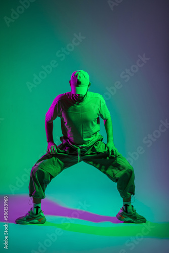 Cool creative fashion man dancer with cap in fashionable clothes with sneakers dancing in colored studio with cyan and magenta light