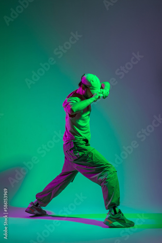 Fashionable handsome professional dancer man in fashion clothes dancing and adjusting his cap in a creative studio with cyan and magenta light