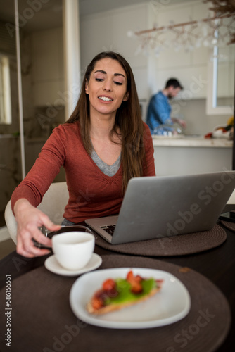 Lovely young woman working from home  while her husband is in the kitchen.
