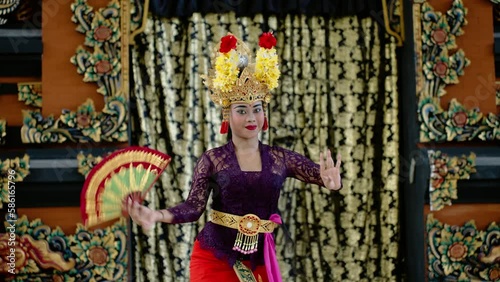 Female dancer in colorful bali national dress with bright makeup moves to sound of musical instruments. Indonesian woman with fan and flower headdress dance to traditional balinese music in temple