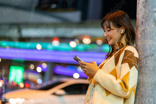 Young Asian woman listening to the music on earphones and mobile phone application during travel in the city at night. Attractive girl have fun outdoor lifestyle using portable device with internet.