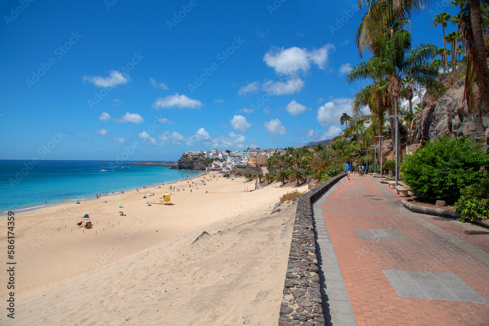 Vista panorámica de la playa de arena blanca y aguas turquesas de Morro Jable en Fuerteventura, desde un paseo peatonal con vistas al resto del pueblo en un día soleado en las Islas Canarias.