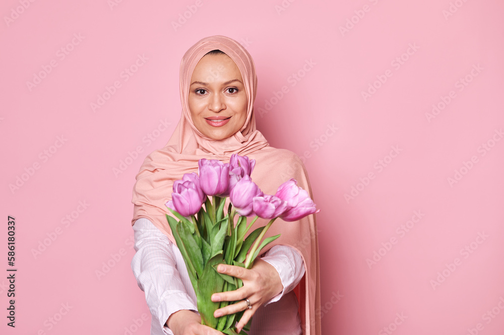 Beautiful happy Muslim woman in pink hijab, holding out at camera a cute bouquet of tulips, smiling looking at camera, isolated over pink background with copy space. Mother's and Women's Day concept