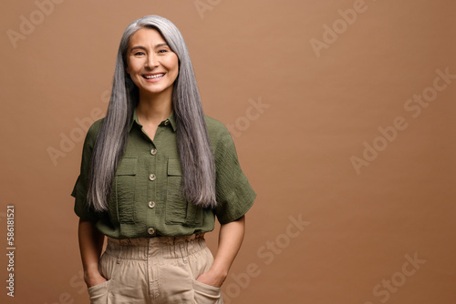 Obraz na plátně Cheerful smiling stylish confident 50 years old Asian beautiful woman standing looking at camera at brown background