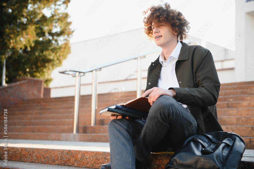 University.Smiling young student man holding a book and a bag on a university background .Young smiling student outdoors Life style.City.Student. University.Smiling young student man holding a book and a bag on a university background .Young smiling student outdoors Life style.City.Student.