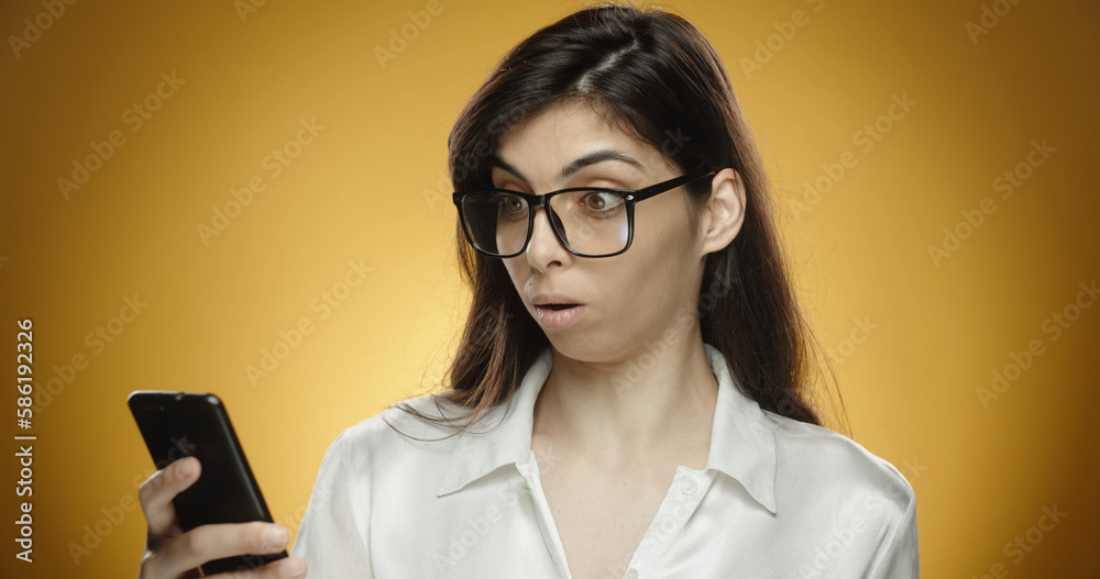 Portrait of a shocked smart businesswoman using mobile phone. Wow and surprise. Woman in studio on a yellow background looking shocked at news. Mobile, social media shock and omg.