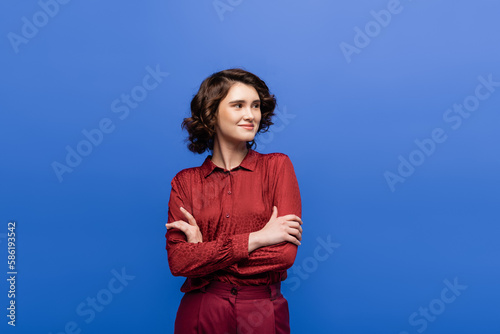 cheerful woman with short curly hair standing with folded arms isolated on blue.
