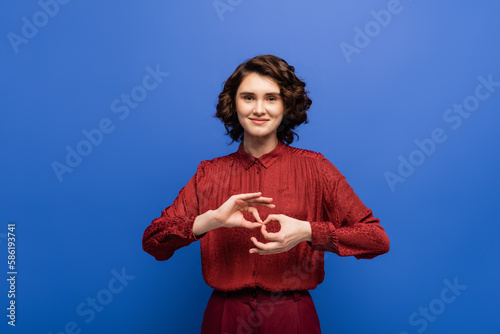 Tableau sur toile young and cheerful woman looking at camera and showing symbol meaning interpreter on sign language isolated on blue