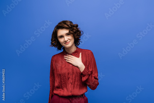 thankful language teacher with short curly hair holding hand on chest isolated on blue.