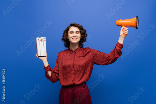 excited language teacher holding megaphone and textbooks while smiling at camera isolated on blue.