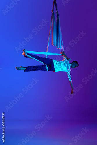 Young, strong, athletic man, professional aerial gymnast training with aerial silk ribbons against gradient blue purple background in neon light. Concept of art, sportive lifestyle, hobby, action and