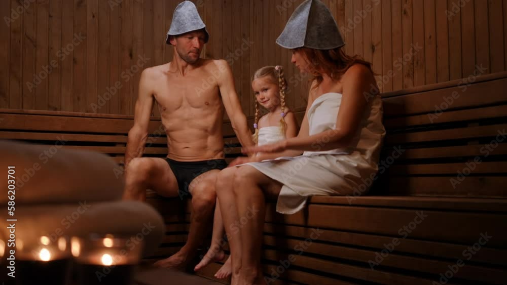 Wide shot carefree daughter with joyful father and mother in Finn sauna indoors. Positive Caucasian man woman and girl talking tickling child laughing. Relaxation and leisure concept