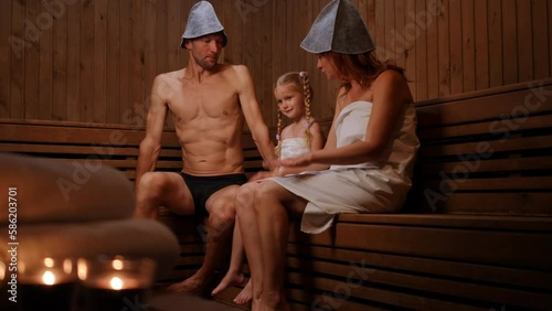 Wide shot carefree daughter with joyful father and mother in Finn sauna indoors. Positive Caucasian man woman and girl talking tickling child laughing. Relaxation and leisure concept